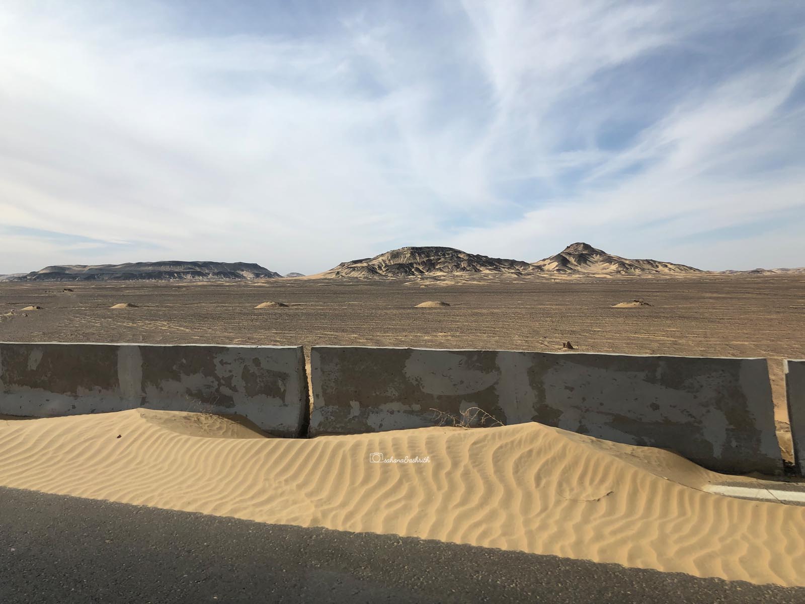 Black short mountains in brown desert with sand touching the side walls of a highway road