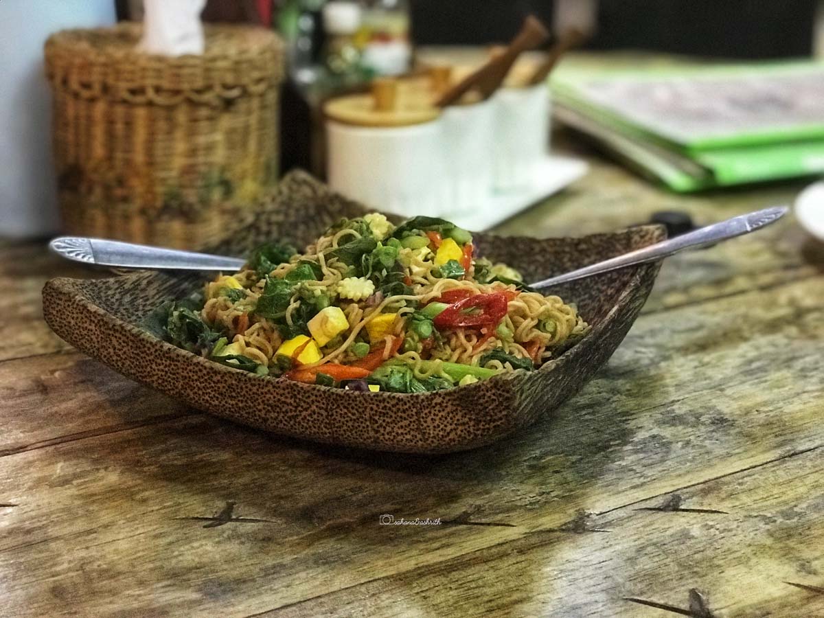 squarish curved wooden plate with noodles topped with tofu, corn, green leaves and a lot of other colourful veggies