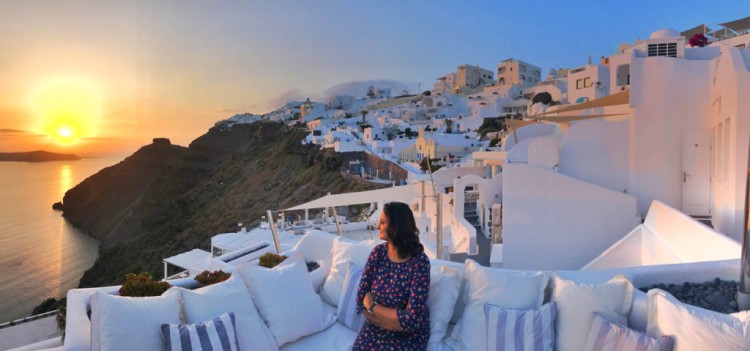 Indian girl watching sunset behind cluster of white houses along the slope of volcanic black mountain by the sea at Firostefani