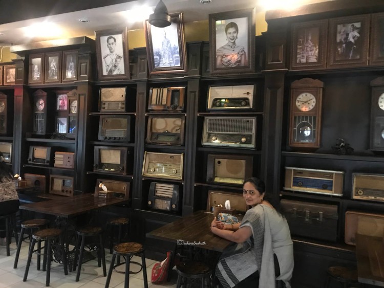 indian woman in grey salwar kameez sitting inside an old cafe in old phuket with antique radios and television sets on the wall