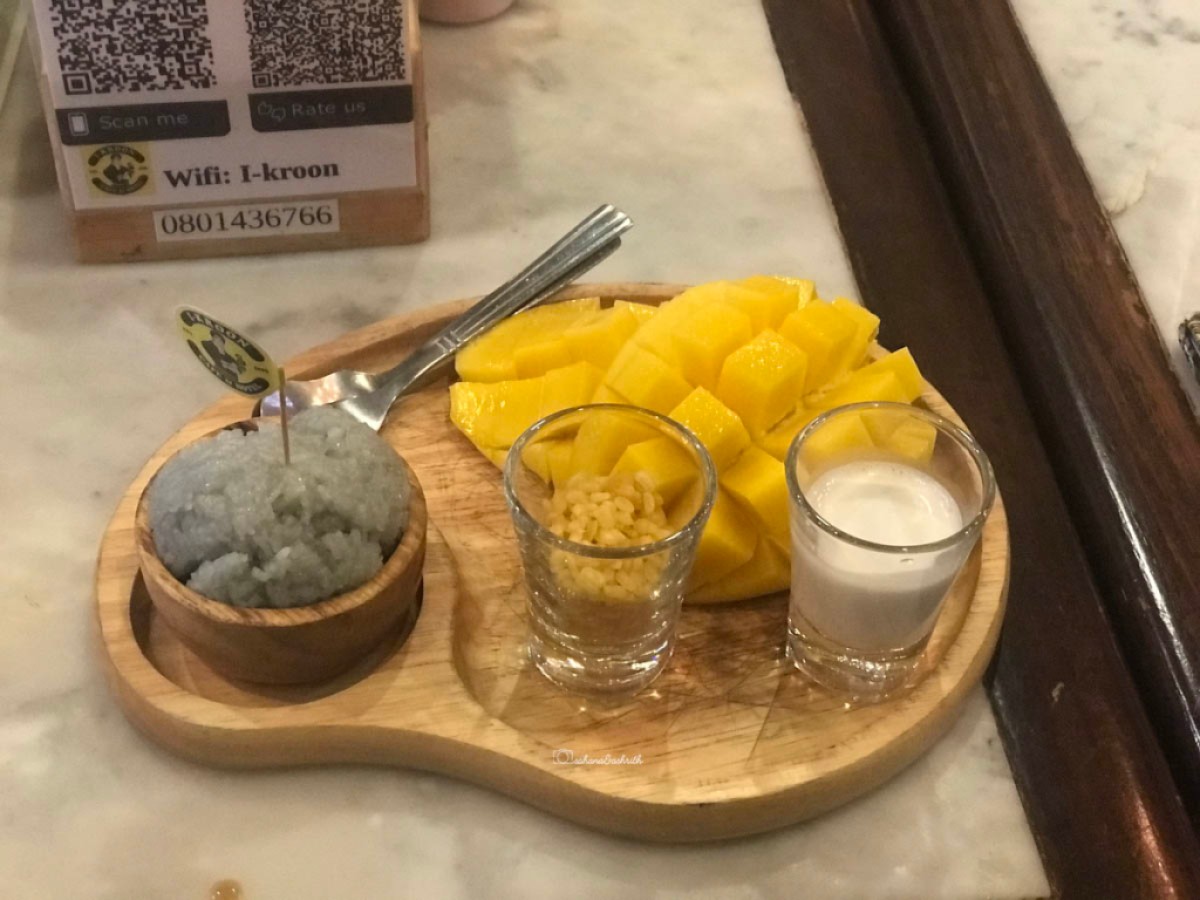mango sticky rice on a wooden plate with grey colour chunk of rice, mango cut into pieces and small glass of coconut cream