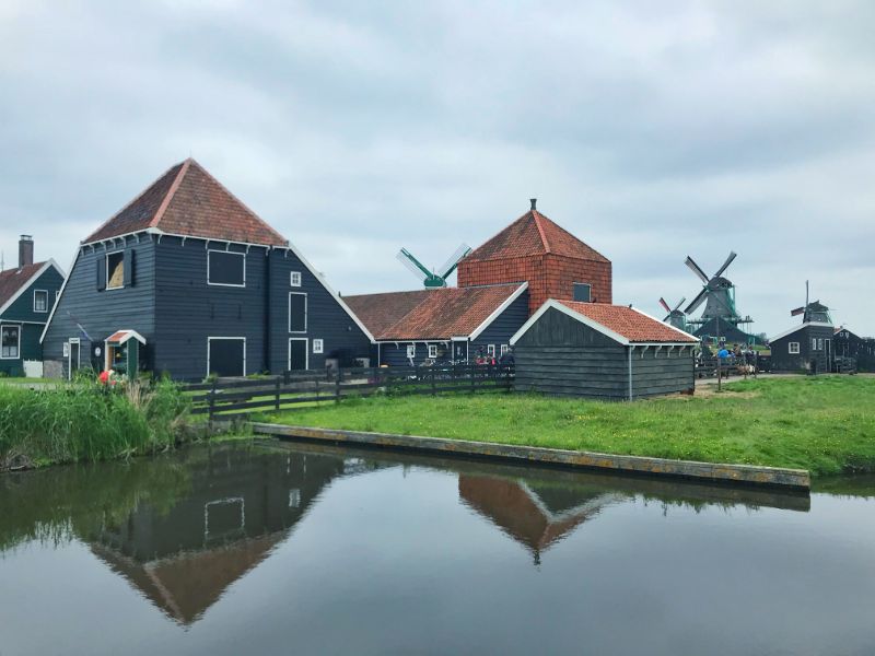 Pyramidical roof over dark green wooden houses by windmill side and their reflextion in water at Znse Sschans- Netherlands itinerary