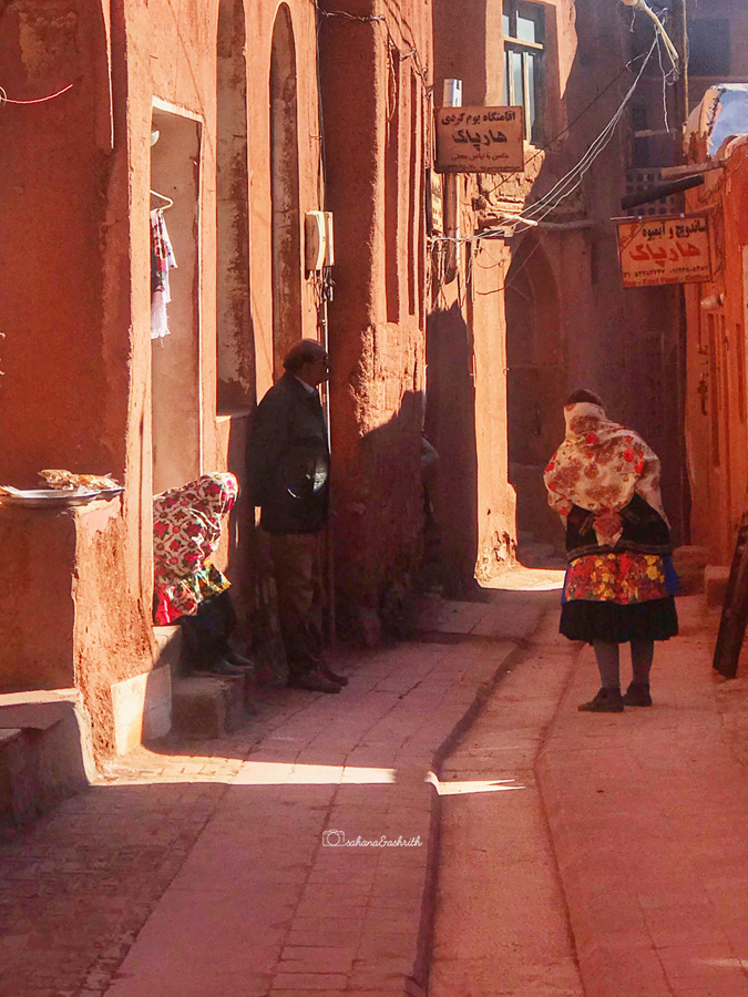 Two women and a man sitting in the shaded allleys of Abyaneh village wearing traditional Shaliteh skirts