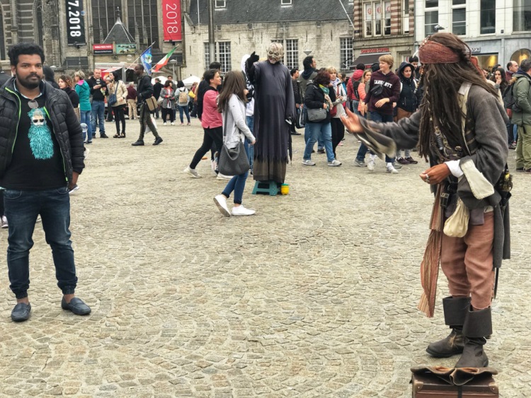 Indian flashpacker at crowded Dam square standing beside artist dressed as Jack sparrow at Amsterdam in the Netherlands