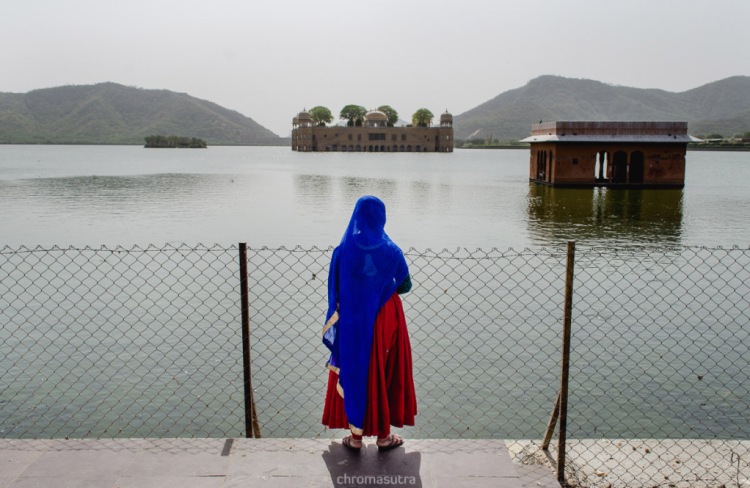 Traditional Rajasthani woman in red Gaghra choli, blue scarf on head standing in front of Udaipur lake